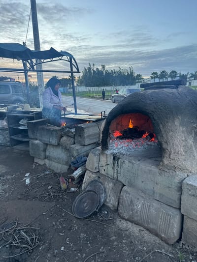 A woman tends a fire in front of a large, stone oven outdoors. The oven is constructed from rough-hewn stone and has a fire burning inside. The woman wears a pink shirt and stands near a metal structure with what appears to be cooking equipment. A car and figures are visible in the background, along with trees and a cloudy sky. The ground is dirt and debris.