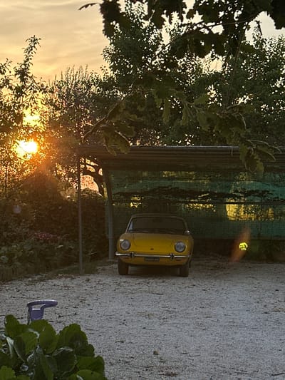 The image depicts a yellow vintage car parked under a covered structure in what appears to be a residential area in Cannara, Italy. The car is facing the camera, positioned centrally in the frame. Behind the car, the covered structure is lined with a green mesh material, and sunlight filters through it, creating a dappled effect. The surrounding environment is lush with vegetation, including trees and bushes, with the sun setting in the background, casting a warm, orange glow. The ground is covered in gravel. A small, purple watering can is visible in the lower left corner of the image, near some green foliage. The overall scene suggests a quiet, rural setting bathed in the soft light of dusk.