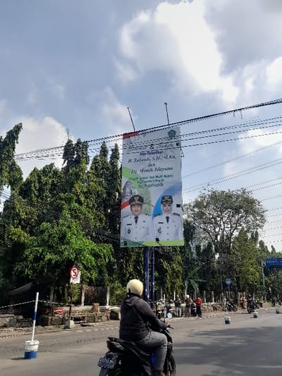 The image shows a street scene in Kecamatan Waru, Indonesia, with a large billboard featuring two people in uniform. The billboard is located on the side of a road with a man on a motorcycle driving past it. The man is wearing a yellow helmet and a black jacket. There are multiple electrical wires stretched across the road. The billboard features a man and a woman, both wearing white and green uniforms and bearing badges. The text above them reads "Selamat & Sukses H. Subandi, S.H., M.M., K.a., dan Hj. Mimik Idayana," suggesting a political or social event. Trees are visible in the background, and the sky is blue with some clouds. The scene appears to be taken during the daytime.