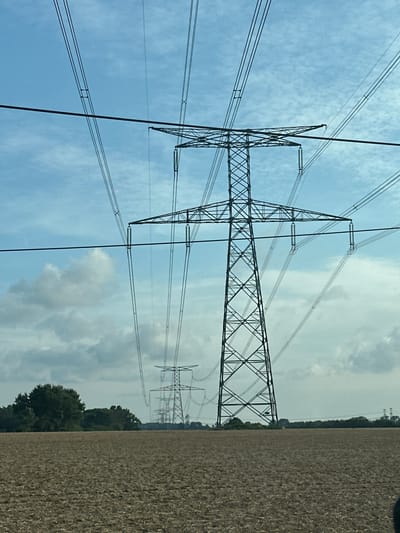 This image depicts a landscape in Varneville-Bretteville, France, characterized by a row of high-voltage electricity pylons stretching across a field under a partly cloudy sky. The foreground is occupied by a plowed agricultural field, showing textures of turned earth and stubble. Behind the field, a line of imposing metal pylons, with their intricate lattice structures, are visible, each supporting multiple power lines that crisscross the frame. The pylons diminish in size and clarity towards the horizon, suggesting a significant distance between them. In the background, a line of trees and foliage separates the field from the distant sky, where scattered clouds break up the expanse of blue. The scene is devoid of any human presence or activity.