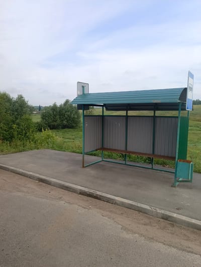 The image depicts an outdoor bus stop in Akulovo, Russia. The bus stop features a metal frame structure with a green, corrugated metal roof. The back wall of the shelter is constructed of grey corrugated metal panels, and a wooden bench is installed for seating. To the right of the shelter, there is a green metal trash receptacle. A blue and white sign with a bus icon is visible on the right side of the structure. The bus stop is situated next to an asphalt road with a concrete curb. In the background, there are green trees and grassy fields, suggesting a rural or semi-rural setting. The sky is partly cloudy with a pale blue hue. No people or vehicles are present in the image.
