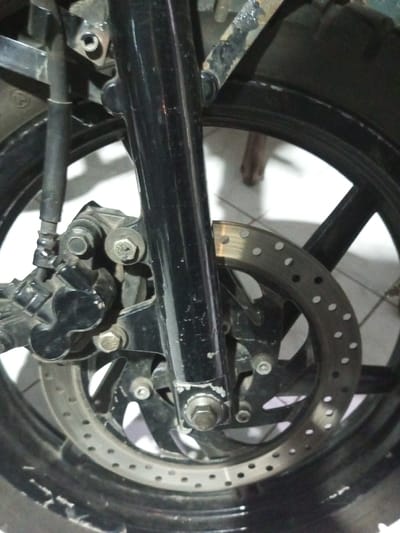 A close-up shows the front wheel assembly of a motorcycle. The black tire and rim surround a perforated brake disc. A black caliper is mounted on the left side, connected by a brake line. Bolts and nuts secure the assembly to the front forks, which are also black. The background features white tiled flooring.