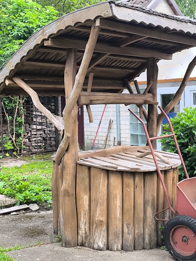 A wooden well structure, made of logs and branches, stands in an outdoor setting. The well has a sloped, shingled roof supported by rustic wooden posts. A red wheelbarrow is positioned to the right of the well, with its wheel visible. In the background, a stack of firewood is enclosed by a metal grate. Green foliage and grass surround the structure, and a building with white brick walls and windows is visible behind the well. The ground appears to be a mix of gravel and concrete. The scene is likely a backyard or garden area.