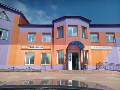 The image shows a two-story building with a distinctive orange and purple facade. The building appears to be a commercial establishment, with signs indicating "STIHL PEAA - Мастер STIHL" on the left side and "КАНЦЕЛЯРИЯ все для школы и офиса" on the right side. The entrance to the building is in the center, featuring a blue awning, a dark metal gate, and double glass doors, one of which is open. A paved walkway with brick patterns leads to the entrance, and a decorative metal railing lines a ramp to the right of the entrance. The building is set against a clear blue sky with scattered white clouds. In the foreground, the reflection of the building and the sky is visible on the hood of a dark-colored vehicle. The scene is likely in Aldan, Russia, a city known for its mining industry and located in the Sakha Republic.