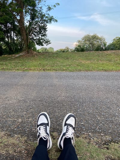 The image shows a person's feet wearing black pants and black and white sneakers. They are standing on a paved path next to a grassy area with trees and vegetation. The sky is blue with some clouds. The sun appears to be out, suggesting it is daytime. The scene is likely an outdoor recreational area like a park or trail. There are no visible text or specific landmarks to identify the exact location beyond what is visually apparent in the natural surroundings.