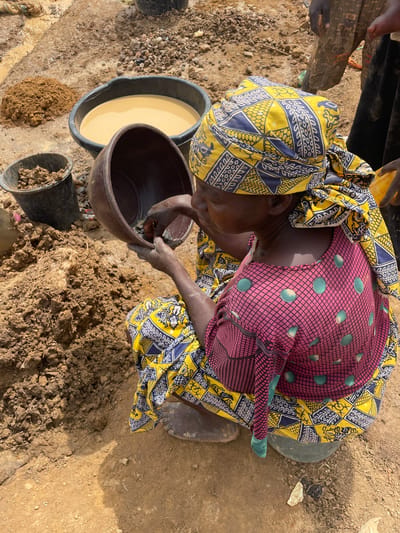 A woman wearing a yellow and navy blue patterned headscarf and a maroon, net-patterned top with green polka dots is kneeling on the ground in Maiyanga, Nigeria. She is interacting with a brown bowl, immersing her hands inside. Next to her is a black bucket filled with liquid and another black bucket filled with dirt. A mound of dirt is on the ground near her. The ground is a mix of tan, brown, and sandy soil.