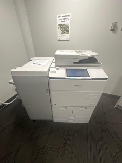 A white Canon imageRUNNER ADVANCE multifunction printer/copier, model P-462, is positioned centrally within an indoor office or service area. The device features a main unit with a central touchscreen display, an automatic document feeder on top, and multiple paper input trays below. A paper finishing unit is attached to its left side, upon which several sheets of paper and a blue pen are placed. The printer rests on a dark gray, subtly textured carpeted floor. The surrounding walls are light gray. A concrete column or pipe is visible in the upper left corner. Directly above the printer, a vertical poster is mounted, displaying the text "This way for all your printing needs." and a graphic depicting a campus map. The poster also includes the text "Clark Center," "Standard for Campus Services Building," and "Canon" and "Epson" brand logos. Two rectangular wall-mounted control panels are visible on the right wall, and a partial view of a white A-frame sign is present on the floor to the right.

The location, indicated by the poster's reference to a "Campus Services Building" and "Clark Center" within the specified city, is likely the Campus Services Building of Wellesley College, Wellesley, United States.