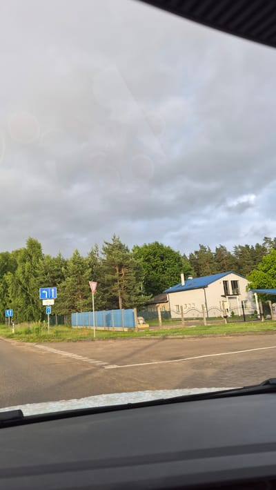 The image depicts a street scene in Vecstropi, Latvia, viewed from inside a vehicle. The foreground is dominated by the dashboard of the car, with a portion of the windshield reflecting a hazy, overcast sky.
The middle ground features a two-lane asphalt road that curves to the left. White dashed lines mark the lanes, and a solid white line indicates the edge of the road. On the left side of the road, there are several road signs. One sign, partially obscured by the windshield reflection, shows two vertical arrows pointing upwards, indicating two lanes ahead. Another sign is blue with white arrows showing lanes going straight and left. A rectangular blue sign with a white 'H' signifies a parking area. A triangular yield sign with a red border and a white center stands near the road.
Beyond the road, a grassy verge separates it from a property. A blue fence runs along the property line, behind which is a white two-story house with a blue roof. The house has dark-framed windows and balconies. A black metal fence encloses the front yard of the house. Mature trees, including a mix of deciduous and coniferous species, form a dense backdrop behind the property and fence. The sky is heavily clouded, suggesting a possibility of rain. The overall lighting is diffused, consistent with an overcast day. There are no visible people or animals in the scene.