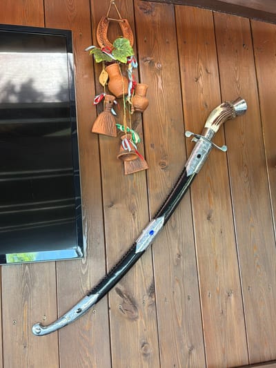 A curved, ornate sword with a decorative silver hilt and a black scabbard adorned with silver filigree and a blue jewel is displayed diagonally on a weathered wooden wall. To the left of the sword, a black flat-screen television is mounted on the wall. Above the television and slightly to the right of it, a collection of clay ornaments hang from twine. These ornaments include a horseshoe, small ceramic jugs, and bell-shaped objects with colorful ribbons. The wall is constructed from vertical wooden planks. The scene appears to be outdoors, likely on a porch or covered patio, given the wood and natural lighting. The location is identified as Nizhnyaya Shilovka, Russia. There are no people visible in the image.