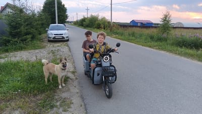 Two young boys are riding on a three-wheeled electric scooter on a paved road in what appears to be a rural or suburban setting, likely Ankudinovo, Russia. The boy in front is driving, wearing a yellow and black tie-dye shirt and patterned shorts. The boy behind him is wearing a brown t-shirt and dark shorts, with glasses. Both boys appear relaxed.

To the left of the road, a light-colored dog with pointy ears is standing on the grass, looking towards the camera. Behind the dog, a silver car is parked on a gravel patch next to a fence. Further behind the car, a wooden house with a dark roof is visible amongst tall evergreen trees.

The road continues into the distance, with fields of grass and wildflowers on either side. In the background, there are several structures, including a blue house with a red roof, a greenhouse, and power lines. The sky is filled with soft, muted colors, suggesting it is either early morning or late evening, with a sunset or sunrise casting a warm glow. The weather appears calm. The visible text on the car's license plate reads "K244MB 199". The scooter has a basket on the front and a storage compartment behind the driver's seat.