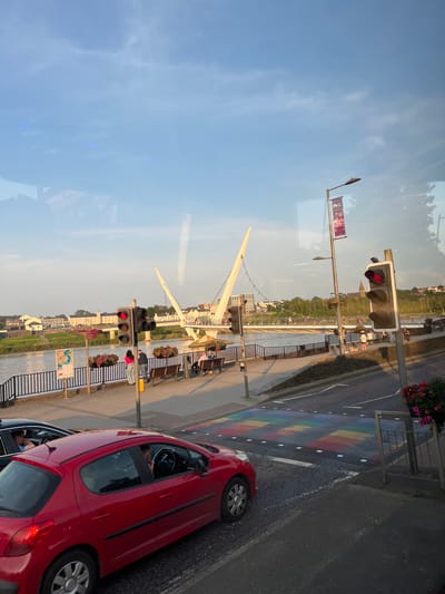 This media file shows an outdoor scene with a river and a pedestrian bridge. The image is captured from a slightly elevated perspective, possibly from a vehicle or a window, and it's oriented vertically.

In the foreground, a red car is parked on the right side of the image, facing away from the viewer. The car's driver-side window is open, and a person is visible inside. Next to the car, a section of road displays a rainbow-colored pedestrian crossing.

The middle ground features a riverbank with people walking and sitting on benches. A distinctive white, modern pedestrian bridge stretches across the river in the background. Traffic lights are visible along the riverbank, with at least one showing a red light.

The background shows buildings on the other side of the river and a treeline. The sky is blue with some wispy clouds, suggesting it is daytime, likely late afternoon or early evening due to the warm lighting. There is a flag with text and a logo hanging from a lamppost near the traffic lights. Another sign with a map is also visible. The scene appears to be in an urban or semi-urban environment, with the rainbow crossing possibly indicating a location known for its inclusivity or a specific event. The overall mood is calm and ordinary, depicting a typical day by the river.