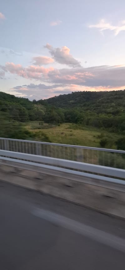 The image captures a view from a moving vehicle, likely on a bridge or overpass, looking out over a lush, hilly landscape. The sky above is a soft gradient of pale blue with scattered clouds tinged with pink and grey, suggesting either dawn or dusk. The hills are densely covered in green trees, with some patches of lighter green, possibly fields or clearings, visible in the valleys. A metallic guardrail runs along the bottom of the frame, and the road surface is blurred due to motion. The scene is evocative of a serene countryside setting, perhaps on the outskirts of Blatino, Bulgaria, where such natural beauty is common.