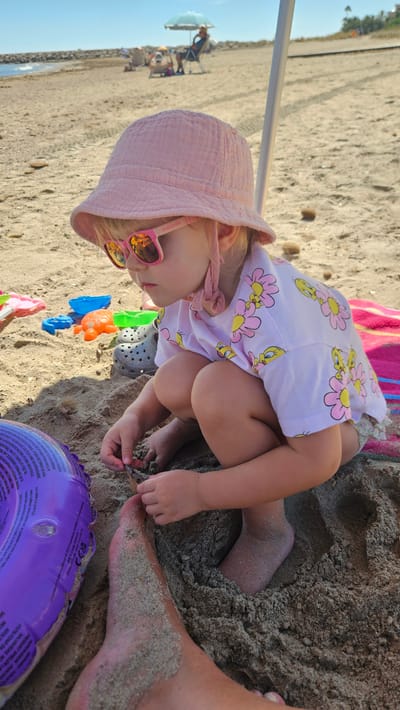In Deveses, Spain, on a sandy beach under a bright sky, a young child with blonde hair is crouched down, engaged in playing with sand. The child wears a pink bucket hat, pink sunglasses with an orange tint, and a light pink t-shirt adorned with a pattern of yellow birds and pink flowers. Nearby, several colorful plastic beach toys are scattered in the sand, including an orange crab-shaped toy and a blue bucket. A gray Croc shoe is also visible. In the foreground, the leg and foot of an adult are partially submerged in the sand, next to a purple inflatable ring. In the background, other beachgoers are present, some sitting under a beach umbrella. The overall scene depicts a relaxed day at the beach.