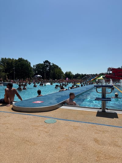 This image captures a lively scene at an outdoor swimming pool on a bright, sunny day. The water is a vibrant blue, and a multitude of people are enjoying the cool water and warm weather. Children and adults are visible throughout the pool, some swimming, others wading, and many simply relaxing.
In the background, a water slide complex adds to the playful atmosphere, with red and green slides visible, suggesting an amusement park or water park setting. The pool area is surrounded by a tan-colored textured surface, demarcated by a blue line. Several trees line the edge of the pool area, providing some natural shade.
The focus of the foreground is on a few individuals near the edge of the pool. One person is partially submerged, looking towards the camera, while another, a man with dark hair, is leaning on the edge, looking out into the pool. The presence of a diving board structure indicates that the pool is equipped for swimming and diving. The overall mood appears to be one of recreation and enjoyment, typical of a summer day at a public pool. The clear blue sky suggests good weather conditions. A small pink ball is visible in the water, hinting at playful activities. A red and white logo is visible on a blue object in the foreground.