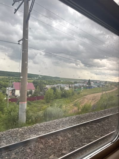 An exterior, eye-level, medium shot captures a view of a rural village from a moving train in Zh/D Razezda Kurkachi, Russia. The foreground features train tracks with ballast, running diagonally from the bottom left to the bottom right. To the left of the tracks is a concrete utility pole with electrical insulators and wires. Beyond the tracks, a landscape unfolds with a cluster of houses, some with red roofs, interspersed with trees and vegetation. A dirt road winds through the village. In the background, rolling hills covered in trees recede towards a cloudy sky. The overall lighting suggests an overcast day.