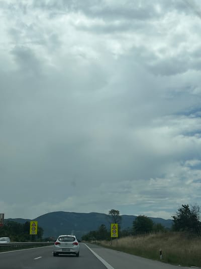 The image captures a highway scene near Slavovitsa, Bulgaria, under an overcast sky. The sky is predominantly grey with extensive cloud cover, showing lighter white patches and minimal hints of blue in the upper right quadrant. The perspective is from a moving vehicle on a multi-lane asphalt road, which features clear white lane markings.
A white Opel Astra hatchback is positioned directly ahead in the right lane, displaying the Bulgarian license plate "CE 4516 AP". A portion of a second white car is visible on the far left, partially obscured by the roadside barrier.
Two identical yellow rectangular road signs are prominent. Each sign features a yellow triangular warning symbol with a black border depicting an upward slope over a horizontal line (commonly indicating a hump or uneven road), and below it, a circular speed limit sign with "100" in black on a white background, denoting a 100 km/h limit. One sign is located on the left side of the highway, and the other further ahead on the right side.
The landscape includes dense green vegetation, comprising trees and bushes, bordering both sides of the highway. Beyond the immediate roadside, a dark, undulating mountain range stretches across the horizon, visible beneath the cloud line. On the right side of the road, a strip of dry, golden-brown grass is visible. No individuals are discernible within the frame.