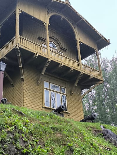 The image shows a two-story wooden building with a distinctive balcony, painted in a muted olive green. The building has a prominent balcony on the upper level, supported by decorative brackets and featuring a wooden balustrade. An arched window is visible on the upper floor, and a large, multi-paned window is on the lower floor. The building is situated on a grassy slope. In the foreground, on the slope, are two old cannons. The overall scene suggests an historical site. The building and cannons are located in Kuldīga, Latvia, specifically at the Kuldīga Castle Mound, a historical defensive structure. The sky is overcast, and there appears to be some precipitation, possibly rain or snow, visible as small white specks in the air. The surrounding trees are lush green, indicating a season of growth.