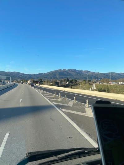 The image depicts a multi-lane highway scene in Le Boulou, France, viewed from inside a vehicle on a clear, sunny day. The foreground shows the vehicle's dashboard, a windshield wiper, and a digital device mounted on the right, displaying a light-colored graphic against a dark background.
The asphalt highway stretches into the distance, with two visible lanes of travel marked by solid and dashed white lines. A dark shadow from the vehicle casting across the left lane. To the right of the main highway, a separation barrier, likely concrete and white, delineates an exit ramp or service road. This adjacent area features black asphalt marked with white chevron patterns and several cylindrical, light-colored bollards along its edge.
Beyond the immediate roadside, green vegetation and some scattered trees are visible. Further in the midground, a "Cédez le Passage" (Yield) sign is identifiable on the right side. Traffic includes multiple passenger vehicles and a prominent white tanker truck traveling in the same direction. In the distant background, a range of mountains with varied tree cover extends across the horizon under a clear, bright blue sky. Some structures and utility poles are visible at the base of the mountains.