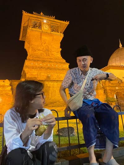 A nighttime wide shot depicts two males in the foreground against the illuminated Great Mosque of Kauman in Kauman, Indonesia. The mosque's prominent square clock tower, lit in a warm orange hue, dominates the left background, featuring a white-faced clock near its top. A golden dome of the mosque is partially visible to the right of the tower. In the midground, a black metal fence separates the subjects from the mosque grounds, with green grass visible behind it.

The male on the right, wearing a black peci, a light-colored short-sleeved patterned shirt, a dark blue sarong with red circular patterns, and light grey slip-on sandals, is seated on the black fence. A light beige sling bag is worn across his chest, and a black smartwatch is on his left wrist. He looks towards the camera with a neutral expression.

The male on the left, with long dark hair and dark-rimmed glasses, wears a white long-sleeved shirt and dark pants/sarong. He is positioned lower than the first male, holding a small, wrapped food item in both hands, and looking towards the male on the right.