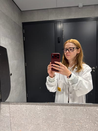 This is a portrait-oriented, medium-shot, mirrored selfie of a young woman in a cleanroom or laboratory setting. She is wearing a white, grid-patterned lab coat with a grey, ribbed collar and cuffs. She has shoulder-length, light brown hair and is wearing black-rimmed glasses. She is holding a dark red smartphone in her right hand, looking at the screen and taking the photo. Her left hand is also visible, adorned with several rings. A black smartwatch is on her left wrist.

The background suggests a modern, sterile environment. Behind her is a large, dark grey cabinet or door with a matte finish and a horizontal handle. To her left, a white wall or door is partially visible, with a round, white sensor or alarm mounted on it. The floor and wall to the right of the cabinet are tiled with a light grey material that has a speckled texture, resembling terrazzo.

The lighting appears to be artificial, consistent with indoor environments. The overall impression is one of professionalism and a focus on cleanliness. A yellow sticker with black text and a symbol is visible on the lab coat, but the text is too blurry to be read. The reflection in the mirror is the primary medium of the image.