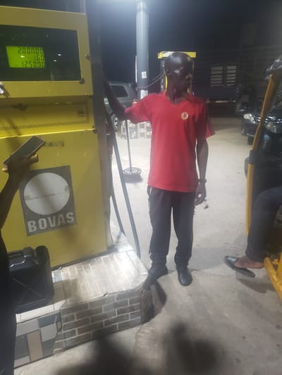 An individual in a red shirt is standing at a yellow gas pump with the logo "BOVAS." The pump indicates a value of "200000.00" and a price of "12500.91." Another individual is holding a smartphone near the base of the pump. A jerrycan sits near the pump's base. The location is in Alalubosa, Nigeria.