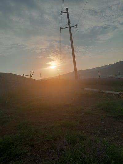 This is a photograph taken at sunset in the city of Кыргыз-Каба, Kyrgyzstan. The image depicts a rural or semi-rural landscape with a prominent utility pole and power lines dominating the center-right of the frame. The sun is low on the horizon, casting a warm, orange glow across the scene and creating lens flare. In the foreground, there is a patch of grass and bare earth. In the middle ground, a fence made of wood and wire mesh is visible, with a rustic wooden structure behind it. The background features rolling hills or mountains, partially obscured by atmospheric haze. The sky is filled with soft, scattered clouds. There are no people or animals visible in the image. The overall impression is one of a tranquil, pastoral setting at dusk.