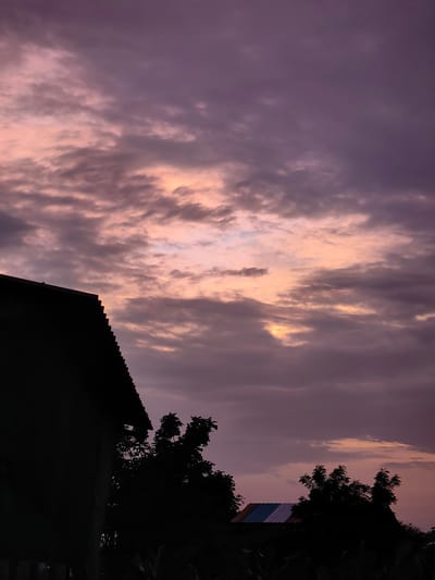 The image captures a twilight sky over Dattapur, India. Silhouetted against the purple and pink hues of the clouds, a portion of a building's corrugated roof is visible in the upper left corner. To the right of the building, several trees are also silhouetted. In the lower right quadrant of the image, another structure with a multi-colored roof can be seen, also in silhouette. The overall impression is of a serene evening scene.