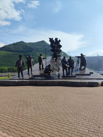 This is a wide-angle, outdoor, eye-level photograph of a large bronze sculpture depicting several figures. The sculpture is situated on a dark, reflective pedestal. In the foreground, a paved area made of interlocking reddish-brown bricks stretches out.
The sculpture features seven human figures in various poses, suggesting collective work or a historical event. One figure stands with an arm raised, gesturing towards the sky, while others are depicted looking at plans, operating equipment, or kneeling. The figures appear to be dressed in practical, older-fashioned clothing. Behind the sculpture, a monumental structure, possibly a dam, with a Russian flag visible, rises from the water. This is flanked by a lush, green, forested mountainside under a bright blue sky with scattered white clouds. The sun appears to be high, indicating it's likely midday or early afternoon. The overall impression is one of grandeur and accomplishment, possibly commemorating a significant engineering or construction project. There is some visible text on the pedestal, but it is not legible in the provided image. The location context indicates this is likely Cheremushki, Russia.
