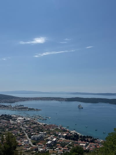 An aerial, high-angle full shot captures a bright, sunny day over a coastal city in Seget Donji, Croatia, with a vast expanse of blue sky occupying the top half of the frame. Scattered, wispy white clouds are visible, with a tiny bird appearing in the distance. The lower half of the image showcases a scenic view of a bustling harbor filled with numerous boats and yachts, and a large cruise ship sailing in the distance. The coastline is lined with a dense cluster of buildings, characterized by red-tiled roofs, and a road winds through the residential areas. The landscape is dotted with lush green trees and vegetation, adding to the picturesque setting.
