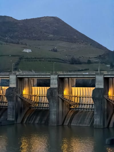The image presents a forensic view of the Carrapatelo Dam, located within the municipality of Lamego, Portugal, during twilight conditions. The lower two-thirds of the frame are dominated by the concrete dam structure, comprising multiple large sluice gates separated by massive piers. The internal mechanisms of these gates are illuminated by warm yellow lights, casting elongated reflections onto the dark, placid water surface in the immediate foreground. Diagonal steel truss structures are visible within the illuminated gate sections. A two-lane roadway, bordered by guardrails and equipped with streetlights, spans the top of the dam. A light-colored sedan is observed traveling from left to right across this road. In the background, a large, dark mountain rises, its slopes exhibiting terraced agricultural patterns, likely vineyards, interspersed with sparse vegetation. A distinctive white, multi-tiered building, identified as Casa Branca de Alvarelhos, is prominently situated on a lower slope of the mountain, slightly left of the image's vertical center. The sky above is a clear, deep blue-grey. No discernible individuals are present in the scene.