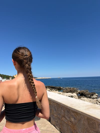 A woman with brown hair in a braid stands facing the sea in Cala Rajada, Spain. She wears a black top and pink shorts. The sea is visible in the background, along with a stone wall and rocky shoreline. The sky is clear and blue.