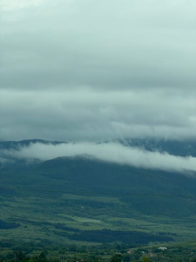 The image depicts a landscape, potentially located near Tedotsminda, Georgia, featuring a mountain range partially obscured by a blanket of low-lying clouds or fog. Lush, green vegetation covers the visible slopes and valleys of the mountain range. The sky above is overcast, contributing to a muted color palette. There are some visible power lines stretching across the image, adding an infrastructural element.