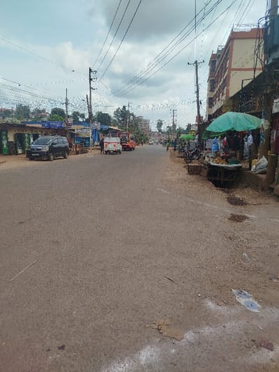 A straight, empty road runs between buildings and shops with pedestrian activity. Electrical lines crisscross overhead. There are some vehicles parked on the left. A green canopy shades a market stall on the right, near a multi-story building. Patches of dirt and minor damage are visible on the road surface. The sky is overcast.