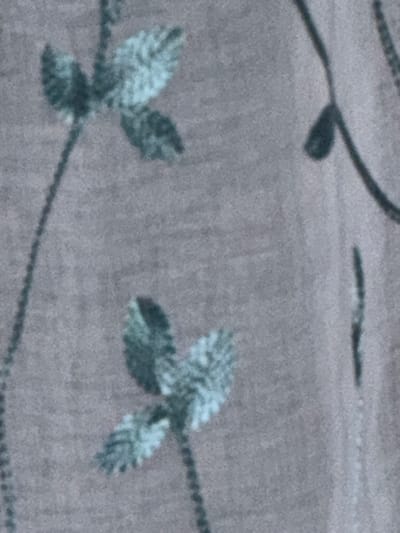 The image displays a close-up, slightly blurry view of a grey fabric with a subtle floral pattern. The pattern consists of stylized leaves and stems rendered in a muted teal or greenish-blue color. There are two distinct floral elements visible. The upper element features two pairs of leaves branching off a central stem, with a lighter color highlighting the texture of the leaves. The lower element is similar, showing two pairs of leaves on a stem, also with a lighter hue suggesting texture. The background is a uniform, textured grey. The image does not depict any elements that would allow for identification of a specific location in Fulda, Germany.