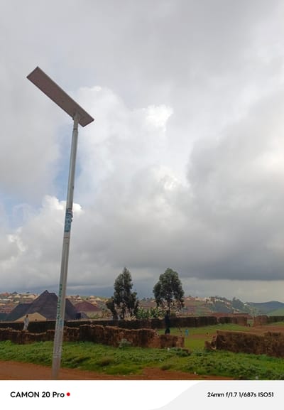 This is a medium shot of a solar-powered streetlamp positioned on the left side of the frame, set against a cloudy sky. The lamp is silver and has a rectangular light fixture. A sticker with a portrait is affixed to the pole. In the background, a settlement with buildings of varying colors and roof types is visible on a hillside. Several trees dot the landscape. In the foreground, there is a low, crumbling brick wall and overgrown green vegetation. A dirt path is in the lower foreground. The image also includes text overlays: "CAMON 20 Pro" in the lower left corner and "24mm f/1.7 1/687s ISO51" in the lower right corner. The location is specified as Nguroje, Nigeria.
