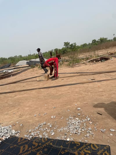 An outdoor scene in Osara Gada, Nigeria, depicts a construction site under a hazy, bright sky. Two adult males are visible on a reddish-brown dirt ground. In the midground, a man dressed in a red tracksuit with white side stripes and open-toed sandals is bent at the waist, facing right, with his hands positioned near a light-colored rectangular wooden block. Several lengths of dark metal rebar rods lie horizontally on the ground in front of him. Behind him and to the left, a second man wearing a gray short-sleeved shirt, dark pants, and a black baseball cap stands, facing right. In the left background, large piles of gray aggregate and additional bundles of rebar are present. Sparse green and dry brown vegetation lines the distant horizon, with some small trees visible. In the immediate foreground-left, portions of black plywood sheets featuring "D.S. HIGH QUALITY" branding in gold lettering are visible. Scattered pieces of gray crushed stone are on the ground in the foreground and midground-right.