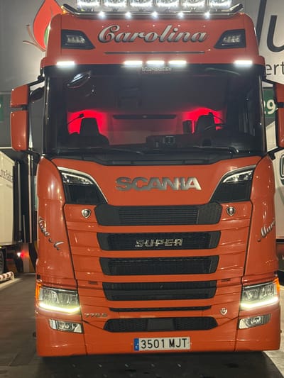 An orange Scania 770S semi-trailer truck cab is centrally positioned, facing the viewer, in a low-light environment in the city of San José Obrero, Spain. The truck's lighting system is active, featuring six circular spotlights on the roof, two horizontal LED light bars above the windshield, illuminated headlights with integrated LED daytime running lights, and fog lights on the lower bumper. The vehicle's interior cab is distinctly lit with red ambient light.

Prominently displayed on the truck's front are the "SCANIA" and "SUPER" badges on the grille, and "V8 POWER" text above the windshield. Custom script text "Carolina" is visible above the windshield, while "Jose A." is written on the driver's side front panel, and "Mario" on the passenger's side front panel. A Spanish license plate, "3501 MJT," is affixed to the lower front bumper.

The truck rests on a paved surface. To the left of the truck, a portion of a white semi-trailer with some dark text is visible. To the right, a structure featuring partial text "ert" and a large numeral "9" is present, alongside a red and orange leaf-shaped logo positioned higher up. No individuals or specific actions are depicted in the scene.
