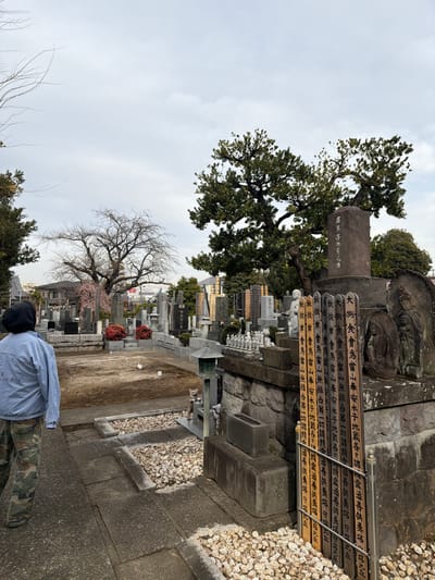 The image depicts a cemetery with various gravestones and memorial markers. The markers are inscribed with Japanese characters. There are also trees, some with dense foliage and others more bare. A person in camouflage pants and a light blue shirt is visible in the foreground. The cemetery has a pathway with gravel and stone features. The overall atmosphere appears serene and respectful.