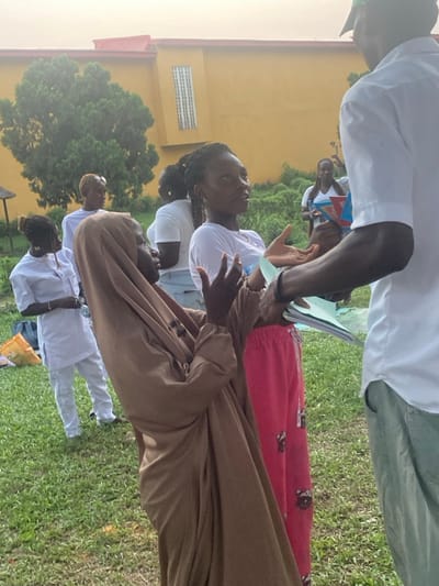 A group gathers outdoors, appearing to be engaged in conversation. Several individuals wear white clothing, including one with a tan hijab. One person wears red pants with a pattern. A man holds papers or a book. A yellow building and trees are visible in the background, and the group stands on a grassy area. The image is rotated.