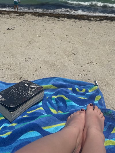 The image captures a beach scene at Hallandale Beach, United States. In the foreground, a blue beach towel with wavy patterns in shades of blue, teal, and lime green is laid on light-colored sand. On the left side of the towel, a paperback copy of the book "HAUNTING ADELINE" by H.D. Carlton, featuring a dark cover with a skull and butterfly design, rests. To the right of the book, a pair of bare feet, with toenails painted a dark red color, are positioned on the towel. The midground shows undisturbed light-colored sand. In the background, the ocean with small waves is visible, separated from the sand by a line of dark seaweed. A lone individual, partially submerged in the water, is visible in the far distance on the left side of the frame. The scene is illuminated by direct sunlight, indicating daytime.