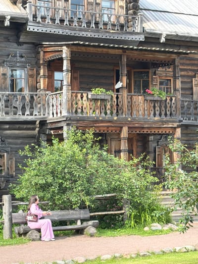 A woman in a pink tracksuit sits on a wooden bench in front of a rustic wooden building. She holds a phone in her right hand and a purse on her lap. The building, with its multiple levels and decorative wooden railings, appears to be of historical or traditional architecture, consistent with a rural or cultural heritage site. Lush green foliage surrounds the bench and partially obscures the lower part of the building, suggesting an outdoor setting with vegetation. The ground in front of the bench is a gravel path. This scene is located in the village of Dnp Ryzhiki, Russia, as indicated by the provided context.