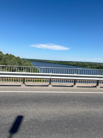 The image shows a view from a bridge over a wide river, likely the Volga, as seen from the city of Yadrin, Russia. The sky is a clear, vibrant blue with a few wispy clouds. A metal railing with vertical bars runs across the foreground, separating the road from the river view. The road surface is asphalt with a white line marking the lane. Lush green trees line the banks of the river, creating a dense, verdant landscape that stretches into the distance. The water is a deep blue and appears calm. The overall scene is one of natural beauty and tranquility.
