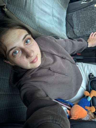 A young woman with light skin and blue eyes is photographed from a high angle, lying down in a vehicle. She is wearing a brown hooded sweatshirt and white pants. Her head is tilted back, and she is looking up at the camera with a slight smile. Her eyes are wide open, and her eyelashes are dark and prominent. A mole is visible on her forehead. The interior of the vehicle is visible, including a gray patterned seat, a black bag, and a stuffed animal. The lighting suggests it is daytime. The image does not provide any information or context to indicate the location is Chernovskoye, Russia.