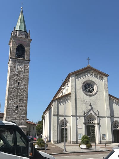 The image depicts a clear, sunny day with a bright blue sky. In the foreground, a white van is partially visible on the left, with its windshield and side mirror showing. To the right of the van, a cobblestone street or plaza leads towards a large church.
The church is a white or light-colored building with a pitched roof. Its facade features a large, circular rose window above a central arched entrance with green double doors. Flanking the central entrance are two smaller arched windows, also with green doors. Decorative architectural elements, including a series of small arches along the roofline and a cross atop the facade, are visible.
To the left of the church stands a tall, stone bell tower. The tower has a square base, progressively narrower upper sections, and a green, conical spire topped with a cross. A clock face is visible on one side of the tower, and an arched opening with a bell inside can be seen higher up.
In the middle ground, several cars are parked on the street behind the plaza. People are walking on the plaza in front of the church, including a woman in a white outfit near a planter box. Small trees and greenery are present in planters on the plaza. The background shows more buildings and trees.
Based on the architectural style, specifically the rose window and the bell tower, this is likely a church in Italy. Given the prompt's request for the location in the city of Udine, Italy, this image appears to be of the Duomo di Udine (also known as the Cattedrale di Santa Maria Annunziata).