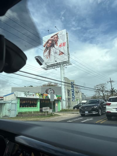 The image shows a street scene in San Rafael, Costa Rica, captured from inside a vehicle. The scene is dominated by a large billboard displaying an advertisement for the clothing retailer H&M. Two women in white clothing are featured on the billboard. The billboard is located at the intersection of two streets, with a streetlight pole supporting it. There are several power lines running across the scene. A building with a green facade and the words "El Rosario" is visible on the left side of the image. The building appears to be a fruit stand or grocery store. On the right side of the image, there is a dark-colored SUV with a license plate that reads "AJ 989" driving on the street. Behind the SUV is a white SUV.  The sky is partly cloudy and the trees are green and lush. There is a sign on the building to the right of the billboard that says "Novaguide."