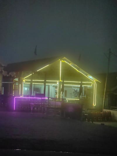 The image shows a building illuminated by yellow and purple neon lights at night in Marhi, India. The building appears to be a restaurant or a similar establishment, with tables and chairs visible in the foreground. The roofline is outlined in yellow lights, and a purple light strip runs along the front of the seating area. The sky is dark and overcast, and there is a utility pole visible to the right of the building. The ground in front of the building is dark and appears to be unpaved. There are no people visible in the image.