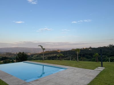 A serene, elevated view captures an infinity swimming pool with bright blue water, bordered by light gray stone tiles and a lush green lawn. Beyond the pool, a rolling landscape unfolds, dotted with trees and buildings, leading to distant hills under a soft, hazy sky with scattered white clouds. Three palm trees are strategically placed on the lawn, adding a tropical touch to the scene. In the far distance, a town sprawls across the valleys and hillsides, illuminated by the warm glow of the setting sun. The overall atmosphere is peaceful and tranquil, indicative of a quiet evening in the countryside near Penafiel, Portugal.