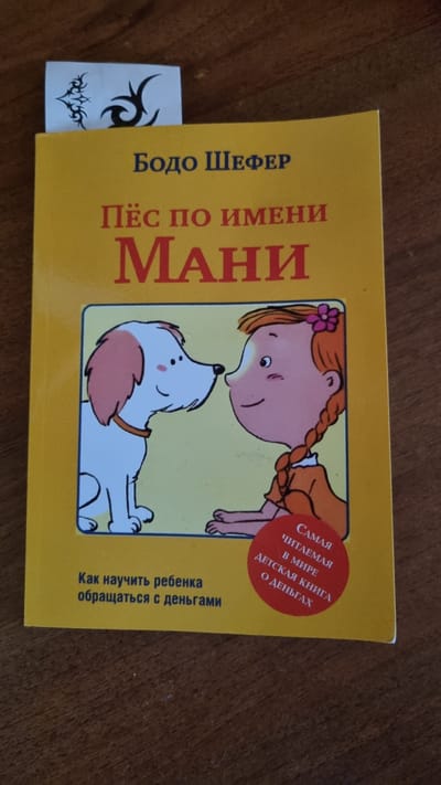 A close-up, overhead shot shows a bright yellow book titled "Пёс по имени Мани" (Dog named Mani) by Бодо Шефер (Bodo Schafer). The book is lying on a wooden surface, likely a table. A white bookmark with a black tribal tattoo design is peeking out from the top of the book. The book cover features an illustration of a happy cartoon dog looking at a girl with braided red hair, who is wearing an orange dress and a pink flower in her hair. Below the illustration, the text reads "Как научить ребенка обращаться с деньгами" (How to teach a child to handle money). A red circular sticker is placed on the lower right side of the book cover, with text that reads "САМАЯ ЧИТАЕМАЯ В МИРЕ ДЕТСКАЯ КНИГА О ДЕНЬГАХ" (The most read children's book about money in the world). The image does not provide any specific information or context related to the city of Солнечная Поляна-3 Снт, Russia.
