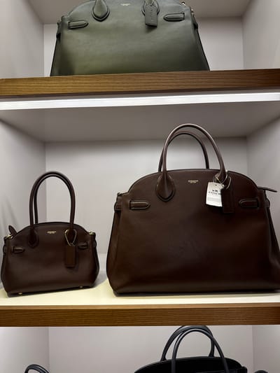 The image shows three leather bags on shelves in what appears to be a retail environment. The bags are positioned side by side and appear to be new with tags. The first bag is a green color, the second one is a large, dark brown bag with a tag on the handle, and beneath it is a smaller version of the dark brown bag. The black bag is cut off and not fully visible on the right side of the image. The shelves have a white background with a wood-like material for the outer frame. Given that the image was captured in Sunrise, United States of America, this is likely a store display in a retail setting.