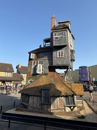 This image captures an outdoor scene with a distinctive, crooked building as the central focus. The building, constructed with dark wooden planks and a terracotta tiled roof, appears to be leaning precariously, with multiple stories and numerous windows of varying sizes. A brick chimney extends from one side. To the right of the building, a purple double-decker bus, labeled "KNIGHT BUS," is parked. A person is visible standing on a railing near the bus.
In the foreground, a group of people are gathered, some seated on bicycles and others standing. They appear to be in a queue, with a sign in the lower right corner bearing the text "SOUND EFFECTS". The scene is bathed in bright sunlight, suggesting a clear day, likely late morning or early afternoon. The sky is a vivid blue. The overall setting evokes a theme park or a movie set, given the theatrical appearance of the building and the presence of the Knight Bus. The architectural style of the building and the bus suggest a connection to the Harry Potter franchise, a popular attraction in the UK. The people appear relaxed and engaged, possibly waiting for an attraction or to take a photograph.